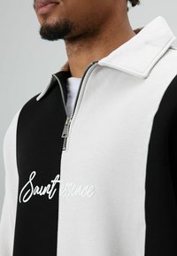 Zwart-witte sweater met rits en kraag, voorzien van een wit geborduurd logo en zachte, gestructureerde stof. Het ontwerp bevat kleurvlakken.