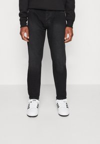 Persona que lleva pantalones ajustados negros y zapatillas Adidas blancas con rayas negras, de pie frente a un fondo claro liso.