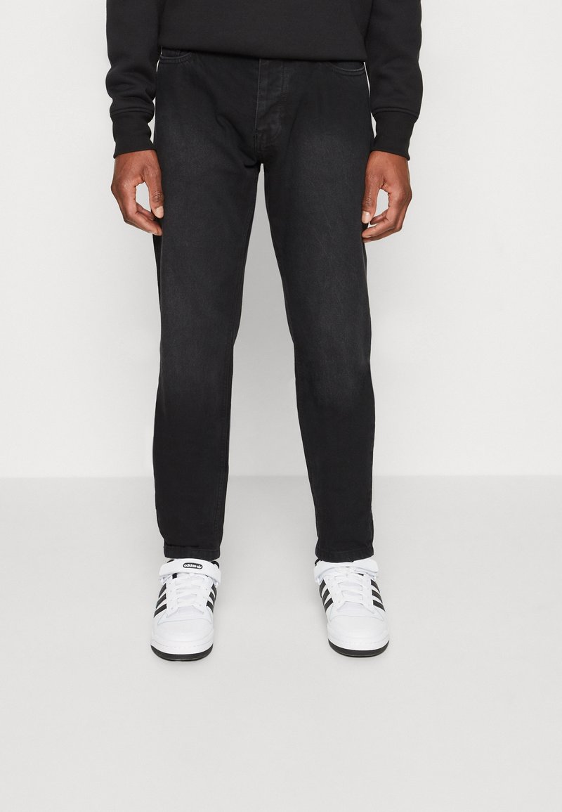 Persona que lleva pantalones ajustados negros y zapatillas Adidas blancas con rayas negras, de pie frente a un fondo claro liso.