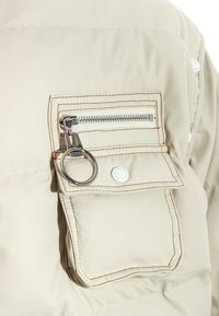 Detalle de una chaqueta acolchada beige que presenta un bolsillo con cremallera con herrajes plateados, costuras en contraste y una pestaña de tirador en forma de anillo circular.