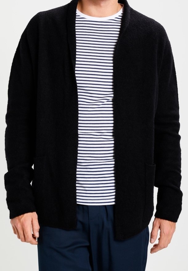 Homme portant un cardigan noir ouvert sur une chemise blanche à rayures bleu marine et un pantalon bleu marine, debout les mains détendues le long du corps.
