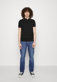 Polo nero con rifiniture rosse e bianche nel collo, design aderente. Abbinato a jeans blu e sneaker nere con suola bianca.
