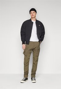 Veste pilote noire, t-shirt blanc, pantalon cargo vert olive avec plusieurs poches, et baskets noires. Couleurs unies et design décontracté.