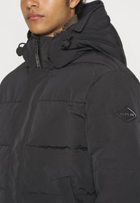 Jachetă neagră padded cu un guler înalt, glugă ajustabilă și buzunare laterale. Dispune de un patch cu logo texturat pe umărul stâng.