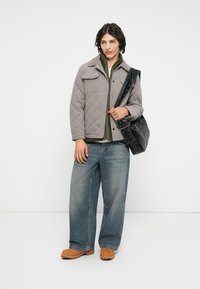 Veste grise matelassée avec des boutons-pression, haut vert en dessous, jean bleu large et sabots marron, tenant un sac en cuir noir.