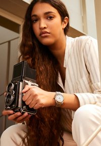 Appareil photo Rollei vintage tenu par une personne avec des cheveux bruns ondulés, portant une chemise blanche à rayures et une montre en argent avec des accents dorés.