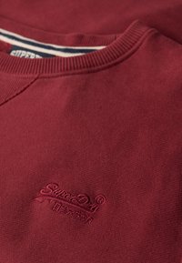 Bordeaux sweatshirt lavet af tekstureret stof, med rund hals og et broderet logo på brystet. Marineblå og hvide striber i den indvendige halsudskæring.