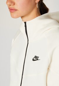 Kremo spalvos džemperis su aukštu apykaklė, juoda užtrauktuku ir juodu Nike logotipu krūtinėje. Pagamintas iš minkštos, tekstūruotos medžiagos.