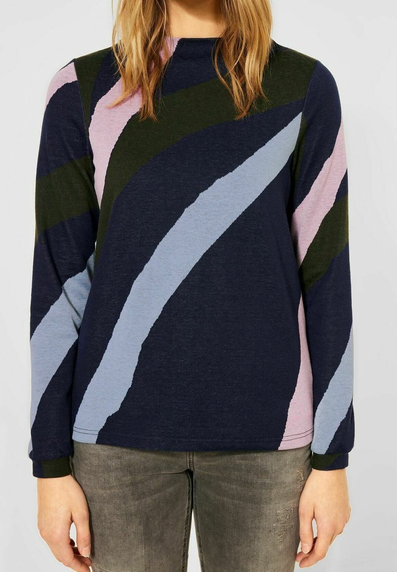 Kobieta w granatowym swetrze z długim rękawem, ozdobionym abstrakcyjnymi, diagonalnymi paskami w jasnoniebieskim, różowym i ciemnozielonym kolorze, w połączeniu z szarymi dżinsami.