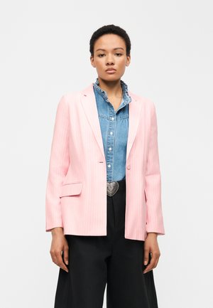 Femme portant un blazer rose clair à fines rayures sur une chemise en jean à volants, un pantalon noir et une boucle de ceinture en forme de cœur, debout devant un fond blanc.