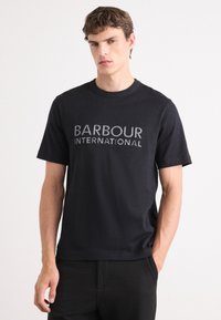 T-shirt de algodão preto com mangas curtas, decote redondo e logotipo "BARBOUR INTERNATIONAL" em fonte branca texturizada na frente.