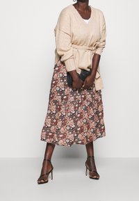 Beige strikket omslagstrøje med bælte, over en blomsterprintet midi-nederdel i sort, pink og hvid. Båret med sorte sandaler med remme.