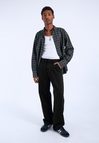 Camisa de cuadros en negro y verde sobre una camiseta sin mangas blanca, combinada con pantalones de pierna ancha negros y zapatillas oscuras. La joyería incluye collares en capas.