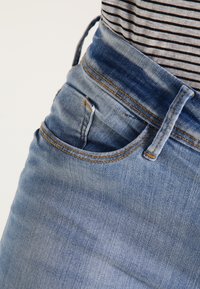 Los jeans de mezclilla azul claro presentan un bolsillo frontal, costuras visibles y una textura ligeramente desgastada. Combinados con una tela a rayas en la parte superior.