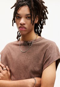 Chemise brune à manches courtes avec un aspect décoloré et un détail en texte noir au niveau du col. Collier en chaîne argentée et bagues portées en accessoires.