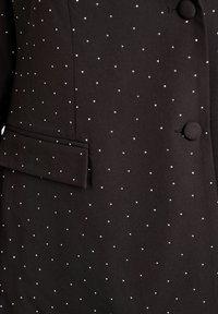 Schwarze Blazerjacke aus Stoff mit kleinen silbernen Nieten, die in einem gleichmäßigen Muster angeordnet sind. Verfügt über eine einzelne Brusttasche und runde Knöpfe.