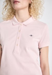 Lys rosa polo skjorte laget av teksturert stoff. Har en klassisk krage, tre knappelukking og et brodert logo på venstre bryst.