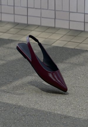 Chaussure plate slingback en cuir verni marron avec bout pointu flottant au-dessus d'un trottoir texturé près d'un mur carrelé.