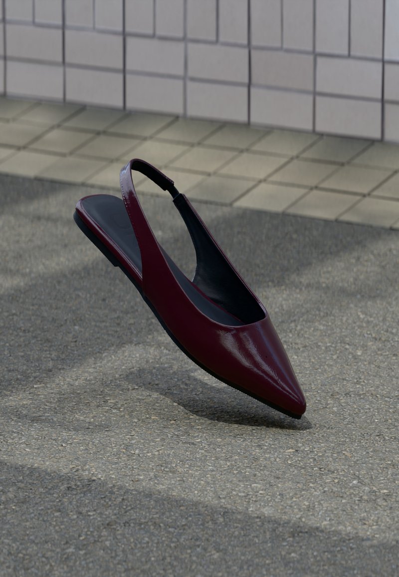 Chaussure plate slingback en cuir verni marron avec bout pointu flottant au-dessus d'un trottoir texturé près d'un mur carrelé.