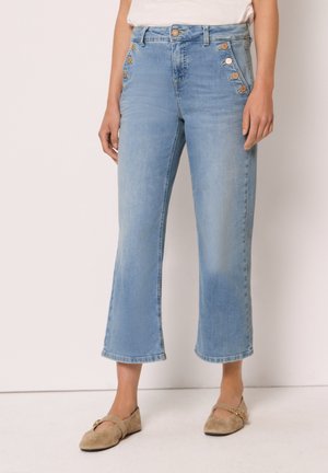 Lyseblå kortere jeans med dekorative gyldne knapper på forlommerne, båret med beige flade sko og en hvid top.