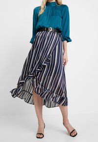Blouse bleu-vert à col haut et détails froncés, associée à une jupe rayée bleu marine avec un ourlet volé et une coupe asymétrique, complétée par des sandales noires.