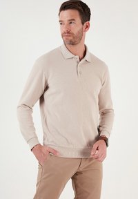Beige hosszú ujjú pólóing bordás textúrával, három gombos lezárással és hemzsebkedéssel, világosbarna nadrággal párosítva.