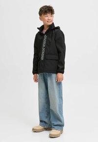 Giacca nera con zip e cappuccio, dotata di tasche frontali, con testo bianco "JACK & JONES". Abbinata a jeans larghi di colore blu chiaro e scarpe beige.