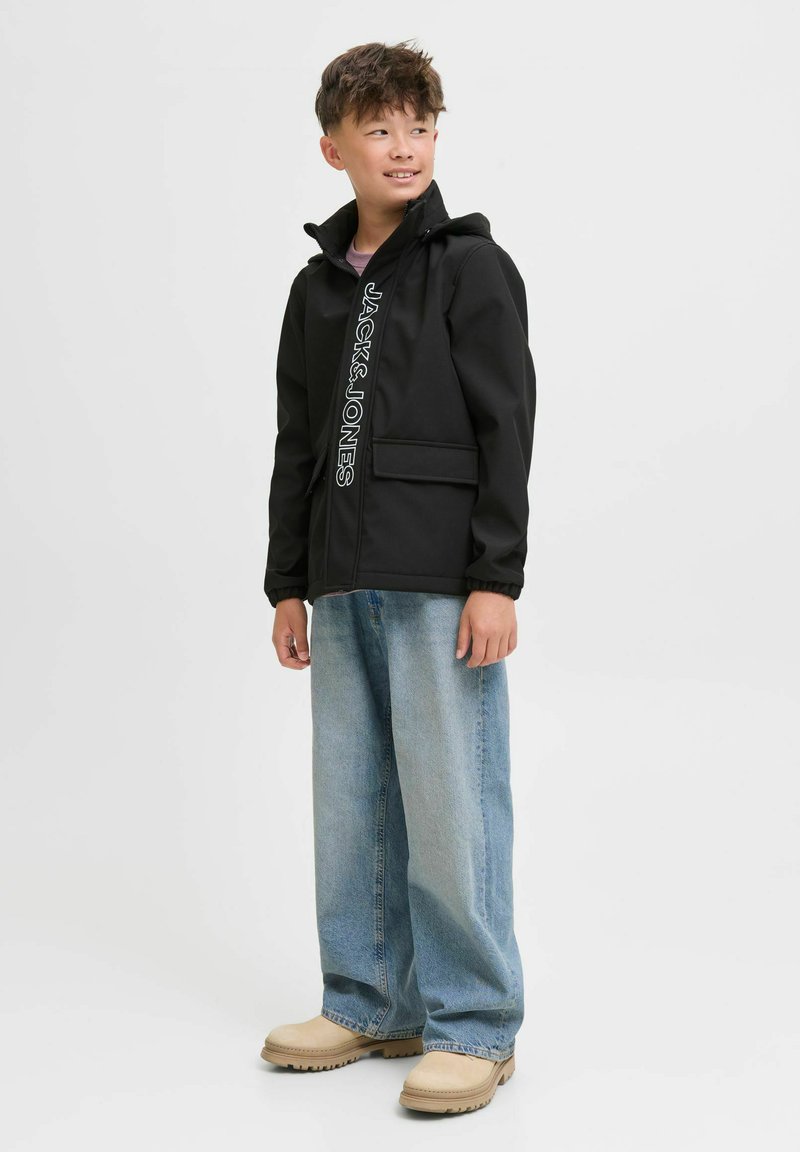Giacca nera con zip e cappuccio, dotata di tasche frontali, con testo bianco "JACK & JONES". Abbinata a jeans larghi di colore blu chiaro e scarpe beige.
