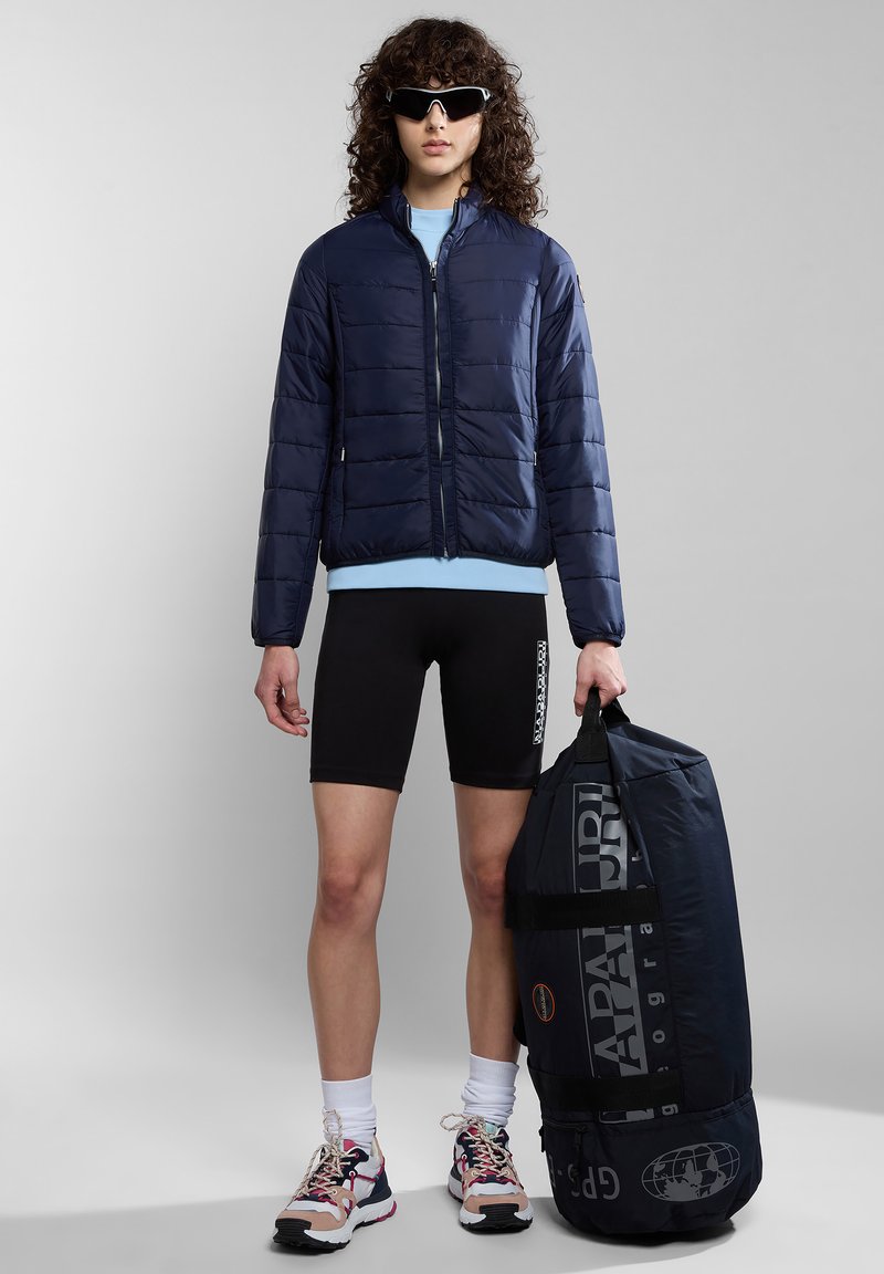 Giacca puffer blu navy con zip frontale, camicia azzurra chiara sotto, pantaloni corti neri da ciclismo e una grande borsa da viaggio blu navy con logo. Sneakers multicolori.