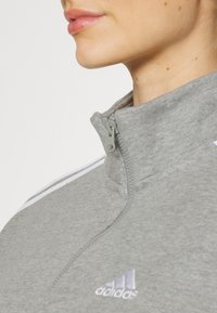 Harmaa vetoketjullinen huppari, jossa on korkea kaulus ja valkoiset raidat hartioilla sekä brodeerattu logo rinnassa. Pehmeä pinta.