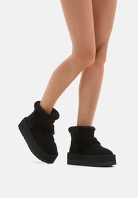 Gambe che indossano stivali alla caviglia neri, con suola spessa e foderati in pelliccia, su uno sfondo bianco, con un piede leggermente sollevato.