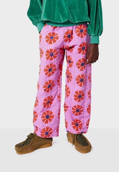 Pantalons matelassés roses avec de grands motifs de fleurs rouges et des centres bleus. Ils présentent des jambes larges et une finition texturée. Associés à des chaussures marron.
