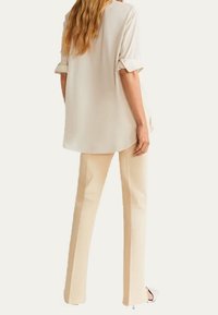 Blusa chiara beige a maniche arrotolate, morbida e scivolata, abbinata a pantaloni affusolati color crema, caratterizzati da una texture liscia e un design minimalista.