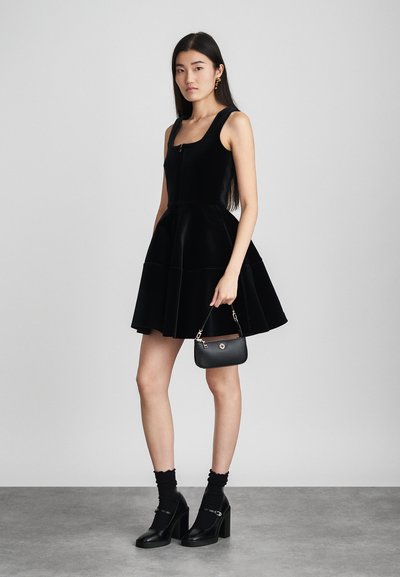 Femme portant une robe noire sans manches et des talons noirs avec des chaussettes, tenant un petit sac à main noir, debout devant un fond gris uni.