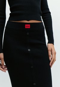 Femme portant une jupe midi côtelée noire avec des boutons sur le devant et une étiquette rouge "Hugo" à la taille, associée à un haut noir à manches longues.