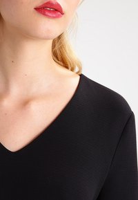 Top nero a scollo a V con tessuto testurizzato, caratterizzato da un taglio pulito attorno al collo e una silhouette liscia. Nessun motivo o accento visibile.