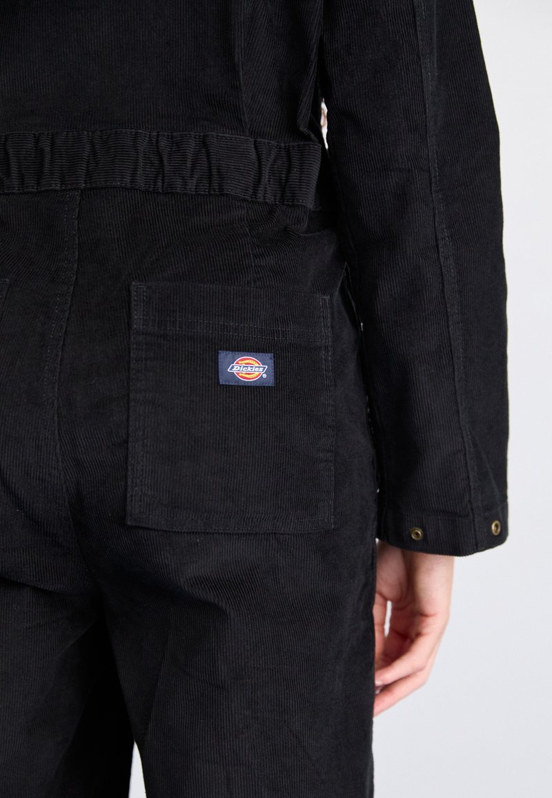 Personne portant un pantalon en velours côtelé noir et une veste, montrant la poche arrière avec le patch logo Dickies sur le pantalon.