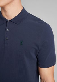 Polo de color azul marino hecho de algodón texturizado, con un cuello clásico, tres botones y un logotipo de ciervo verde bordado en el lado izquierdo del pecho.