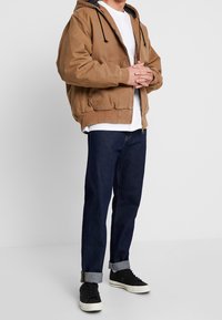 Brun zip-up hoodie med dragsko, bärs över en vit t-shirt, kombinerad med mörka denimjeans och svarta sneakers med vita sulor.