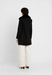 Abrigo negro con cuello de felpa, corte a medida y mangas largas, llevado sobre pantalones de pierna ancha en color beige claro. Presenta líneas limpias y un diseño minimalista.