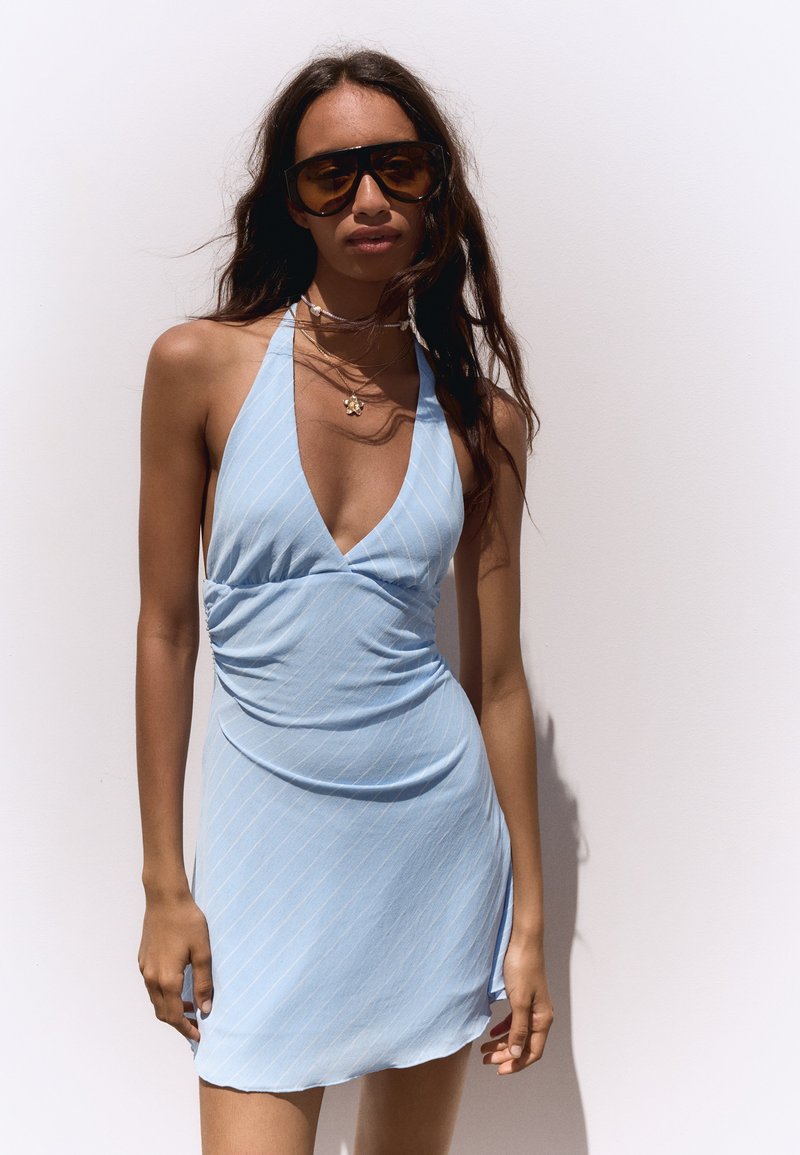Femme portant de grandes lunettes de soleil et une robe bleu clair à col licou avec un décolleté en V profond sur fond blanc.