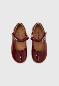 Ζευγάρι γυαλιστερών σκούρων κόκκινων παπουτσιών Mary Jane με κυματοειδείς άκρες και ρυθμιζόμενες λουρίδες με αγκράφα, από πάνω σε απλό φόντο.