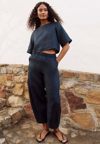 Dark navy blue cropped linen shirt with wide sleeves, paired with loose-fitting matching pants. Model wears black sandals, standing against a stone wall.
