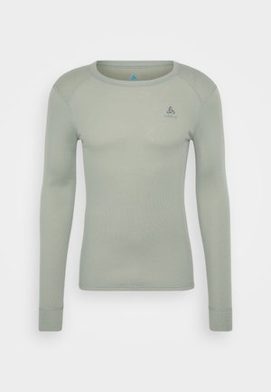 Langermet topp i lys grønn, laget av et teksturert materiale. Har rund hals og en liten grå logo på venstre øvre bryst.