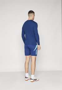 Camiseta de manga larga de color marino y pantalones cortos a juego con toques de azul claro y tres rayas verticales. El modelo lleva zapatillas deportivas blancas con suelas naranjas.