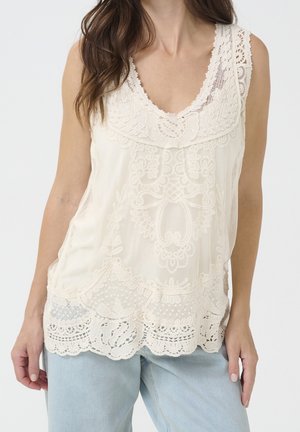 Top de encaje color crema con escote en V, patrones florales intrincados, dobladillo festoneado y ajuste holgado, combinado con jeans azul claro.