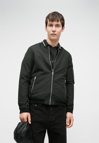 Ung mann i svart zip-up jakke og svarte jeans som holder en svart lærhatt mot en hvit bakgrunn.