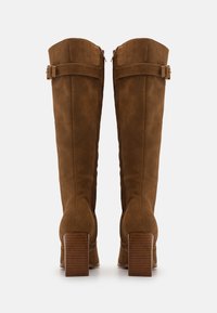 Botas de ante marrón hasta la rodilla con tacones de bloque gruesos, que presentan un detalle de correa ajustable en la parte superior y una textura suave.