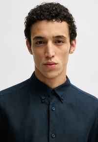 Ciemnoniebieska koszula z kołnierzykiem, z zapięciem na guziki i gładką teksturą. Materiał wydaje się lekki z subtelnym połyskiem.