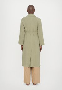 Cappotto lungo verde chiaro con vita cinturata, dotato di colletto a risvolto e maniche lunghe. Indossato sopra pantaloni beige a gamba larga.